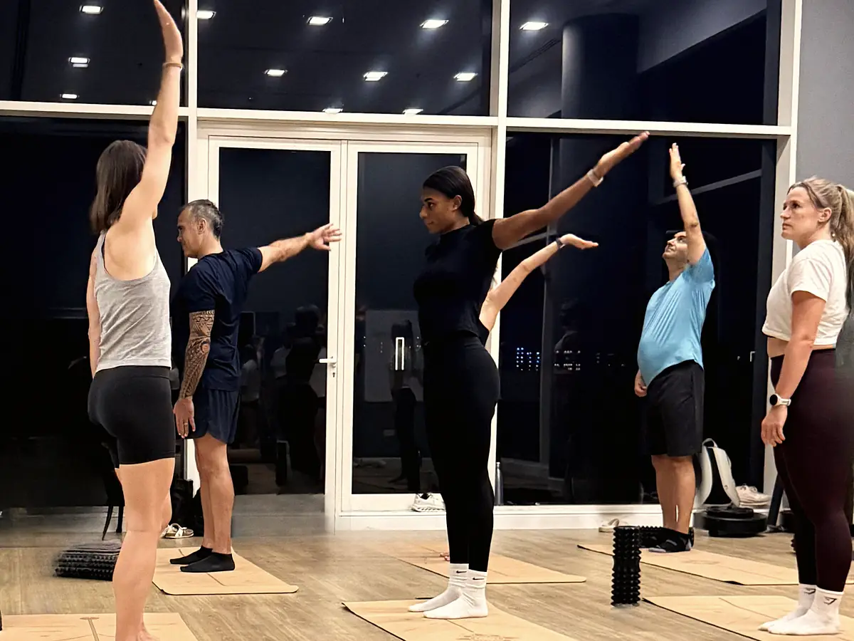The pilot room standing on cork mats, every athlete reaching one arm long overhead